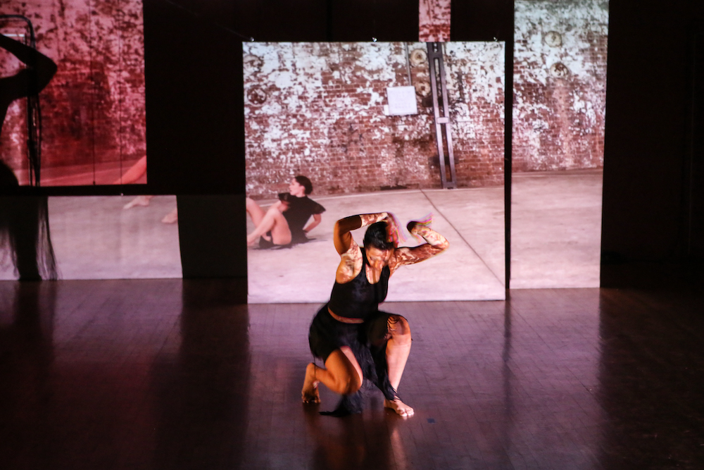 two women dance intensely. one is behind the other in a video projection the other, in front of the projection, hovers above the floor with intense intention.